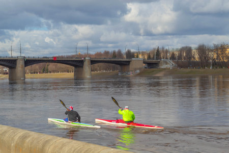 The photo shows the nature and sports life of the city. On the river, two men are engaged in rowing....