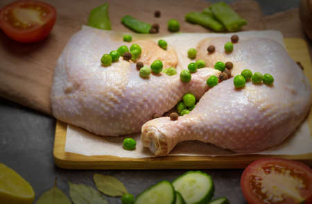 Two raw chicken legs are lying on a bamboo board, and next to them are fresh vegetables and spicesの写真素材