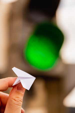 One small paper airplane in hand against a traffic light background with a glowing green signal. Permission to fly. Takeoff is approved and allowed. Vertical shot. Concept.の写真素材