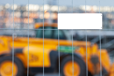 White empty mockup plate with copyspace fixed on a metal grid. Behind the fence in the background is a yellow construction truck. Empty blank sign on blurry background. Advertising conceptの写真素材