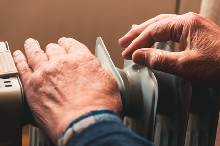 An elderly man warms his hands over an electric heater. In the off-season, central heating is delayed, so people have to buy additional heaters to keep houses warm despite increased electricity bills.の写真素材
