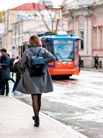 Unrecognizable young blond woman in a short light coat rush to tramway. Black stylish backpack on her back. Female in black stockings walking along the street to the tram stop. Warm sunny autumn dayの写真素材