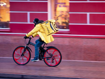 Moscow, Russia - January 17, 2020: Courier of food delivery service from restaurant and cafe Yandex Food rides bicycle against wall of old mansion. Man in branded yellow clothing with thermal backpackのeditorial素材