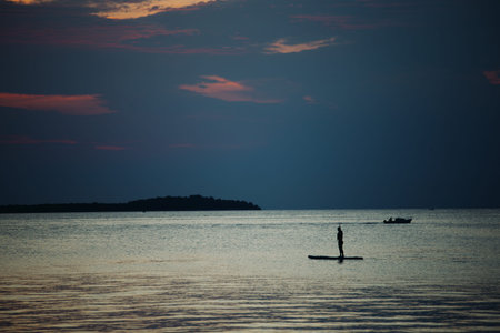 A swimmer stands on a sup board in the middle of the sea on a summer evening at sunset. Light...