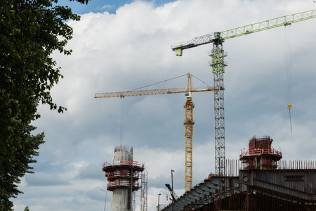 Construction site features multiple cranes and scaffolding under a dramatic cloudy sky, highlighting...