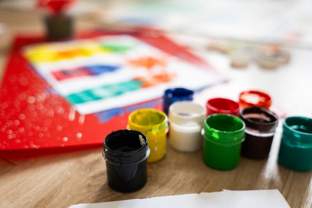 Colorful paint containers are arranged on a wooden table, surrounded by various art supplies and a vibrant artwork in the backgroundの写真素材