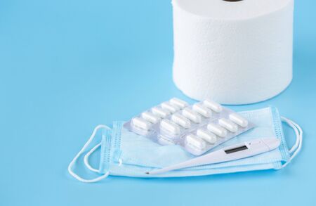 Face mask, thermometer, pills, toilet paper roll on a blue background. Medicine supplies for home coronavirus COVID-19 quarantine. Close up view.の写真素材