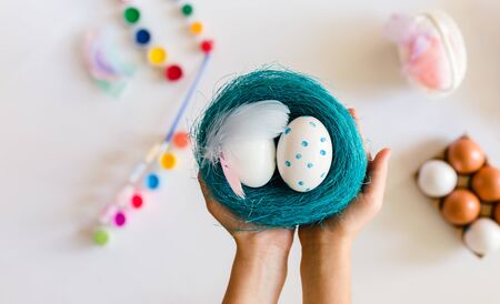 Close up view hands holding blue basket or nest with Easter eggs on white background. Coloring eggs for easter t at home. Happy Easter concept.の写真素材