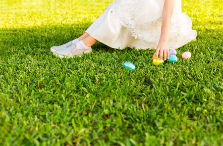 Little girl in a beautiful white dress hunting for easter egg on Easter day. Child having fun outdoor. Kid playing with eggs on green grass. Spring holidays concept. Copy Space for textの写真素材