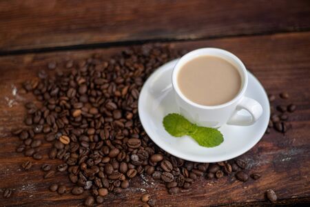 Cup of coffee on a dark background. White milkman. Milk is pouring from it into a cup. On a saucer there are mint leaves, brown grains are scattered nearby,の写真素材