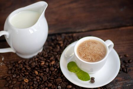 Cup of coffee on a dark background. White milkman. Milk is pouring from it into a cup. On a saucer there are mint leaves, brown grains are scattered nearbyの写真素材