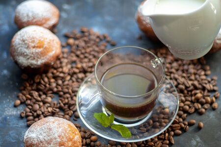 Cup of coffee on a dark background. White milkman. Milk is pouring from it into a cup. On a saucer there are mint leaves, brown grains are scattered nearbyの写真素材