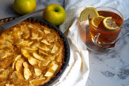 Charlotte apple pie in a round shape, decorated with fan-laced apple slices from the top, a piece of cake next to it, a cup of tea, autumn leaves, all against a dark background.の写真素材