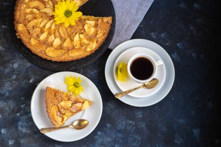 Charlotte apple pie in a round shape, decorated with fan-laden apple slices from the top, a piece of cake next to it, a cup of coffee, autumn leaves, all against a dark background.の写真素材