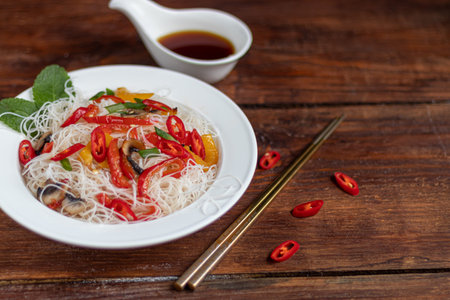 Oriental rice noodles Udon with sweet pepper, mushrooms. On a wooden background. Near chopsticks. A...