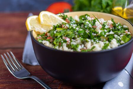 Tabule salad in a round plate on a wooden background. Couscous, parsley with green onions and tomatoes and mint. Dressed with olive oil and lemon juice. Copy space.の写真素材