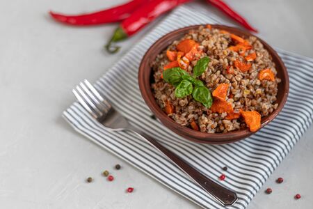 Buckwheat porridge in a bowl with stewed carrots. Decorated with green leaves. On a cotton napkin, on a light gray background under concrete. Copy space.の写真素材