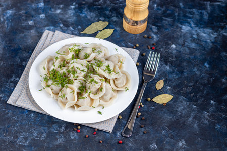 Boiled dumplings on a white plate. In the background is a wooden hodgepodge, greens, red peppers and bay leaves. Dark marbled background. Copy space.の写真素材