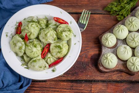 Boiled khinkali made of dough with spinach. Garnished with Red Chilli, Dill and Parsley. On a wooden background. Nearby on a brown board are raw khinkali frozen. Copy space.の写真素材