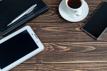 A wooden office desk with many things on it. Diary with pen,  phone. White cup of coffee. Top view...
