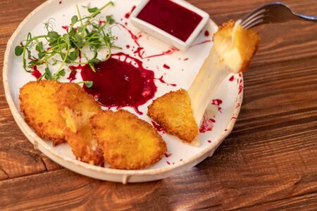 Camembert cheese fried in batter and breadcrumbs with cranberry sauce. Soft and viscous inside. The plate is decorated with a spray of sauce and microgreen. On a wooden background.の写真素材