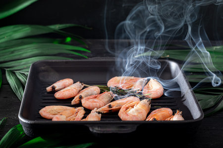 Grilled prawns in a pan with a sprig of rosemary for frying. On the substrate are green palm leaves. On a black wooden background. Copy spaceの写真素材