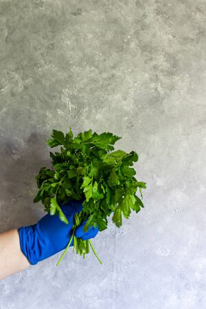 A female hand in a blue disposable glove holds different objects. Spray for disinfection, mandarin and a bunch of greens on a gray background. Copy space.の写真素材