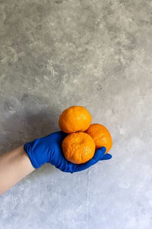 A female hand in a blue disposable glove holds different objects. Spray for disinfection, mandarin and a bunch of greens on a gray background. Copy space.の写真素材