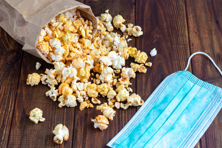 A transparent bowl of popcorn on a wooden surface. Next to a laptop and headphones. The mask...