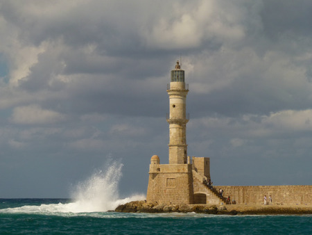 Gloomy gray clouds hang over the lighthouse of yellow and white bricks in Chania, about which waves crash, exploding in a cascade of splashes, Creteの写真素材