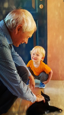 The little two-year-old boy happily smiles and looks at his grandfather with adoration, who strokes a black and white cat on a summer dayの写真素材