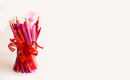A bunch of sharpened pencils, felt-tip pens in red and pink, and a pen in the shape of a bunny, tied with a scarlet ribbon on a white background with space for copying, pasting text, and inscriptionsの写真素材
