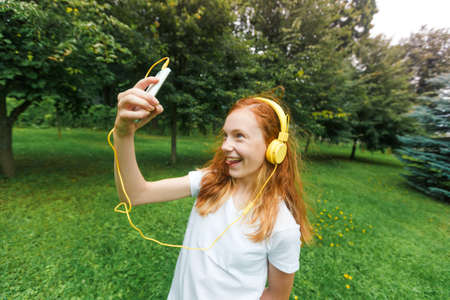 red-haired girl listens to lessons in yellow headphones. Teenager in white t-shirt and headphones in the park,の写真素材