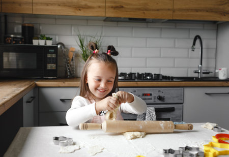 a cute little girl in an apron is sitting at the kitchen table and playing with the dough.