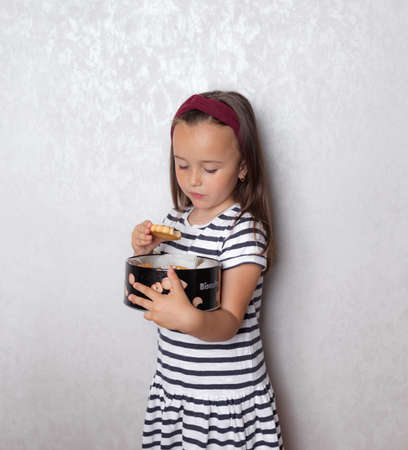 a little girl in a striped t shirt with black and white stripes is standing against the wall eating cookies from a large boxの写真素材