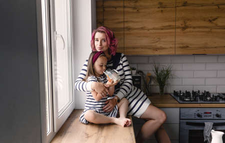 happy family, mother and her little daughter are sitting on the windowsill in the kitchen and drinking milk. They are dressed in beautiful dresses with black and white stripes.の写真素材