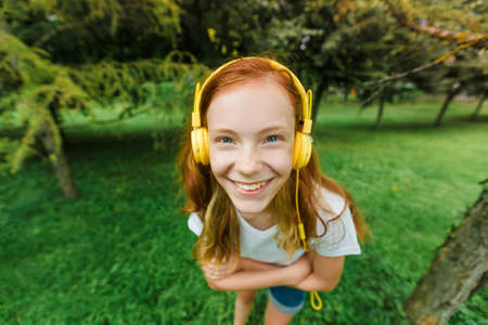 a teenage girl with red hair listens to music with headphones in a Park in the summer. a young woman enjoys and relaxes outdoors on a Sunny day.の写真素材