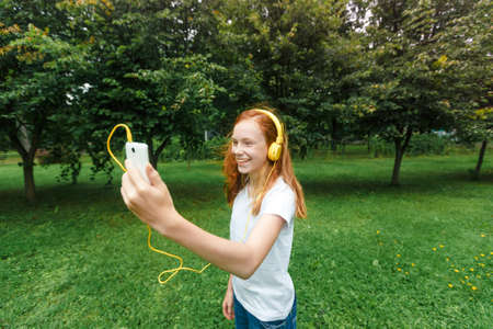 a teenage girl with red hair listens to music with headphones in a Park in the summer. a young woman enjoys and relaxes outdoors on a Sunny day.の写真素材