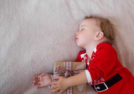 a little girl is sleeping on a bed in a Santa Claus costume and hugging a Christmas gift.