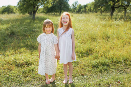 two little girls sisters play and have fun on the grass holding hands in light dresses.Summer, Sunny weather in a good mood.の写真素材