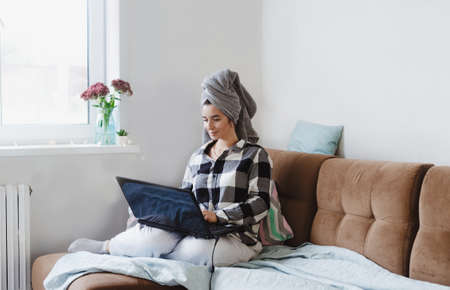 A relaxed woman in a plaid shirt with a towel on her head is working on a laptop doing online shopping, checking social networks, reading news or chatting on the Internet, sitting on the couch. Hotel room.の写真素材