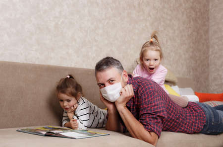 A young father at home in self-isolation reads a book to his beloved daughters. Work at home.の写真素材
