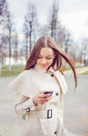 a beautiful young girl in a light raincoat dials a number on the phone.の写真素材