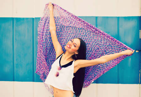 a beautiful young girl in a white t-shirt and blue jeans holds a large multicolored handkerchief at arm's length against a blue wallの写真素材