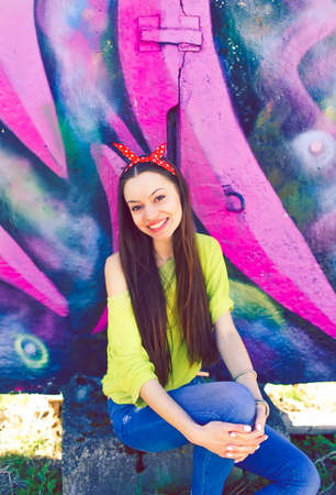 A stylish teenage girl in a yellow sweater and blue jeans poses near a graffiti wallの写真素材