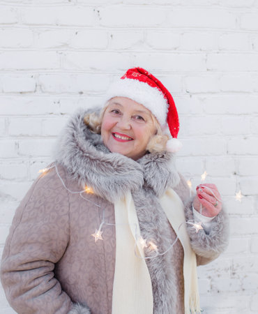 Portrait of an elderly woman in a red Santa hat on the background of the wall. An old woman in a winter jacket with lights in her hands on an isolated white brick background.の写真素材