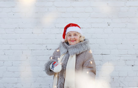 Portrait of an elderly woman in a red Santa hat on the background of the wall. An old woman in a winter jacket with lights in her hands on an isolated white brick background.の写真素材