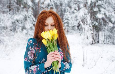 A girl with red hair holds a bouquet of yellow tulips on a winter background. Flowers as a gift for womens Day. March 8.A beautiful girl smiles.の写真素材