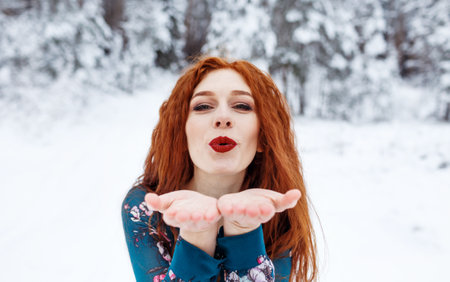 Young red-haired girl with long red hair on a winter background.Air kiss. Red-haired woman in a summer dress on the background of winter nature.の写真素材