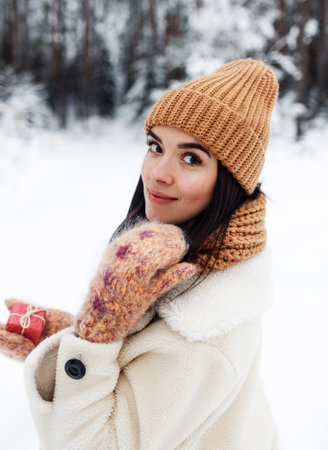 A beautiful happy laughing young girl in gloves and a winter hat-scarf, covered with snow flakes. Winter forest landscape backgroundの写真素材